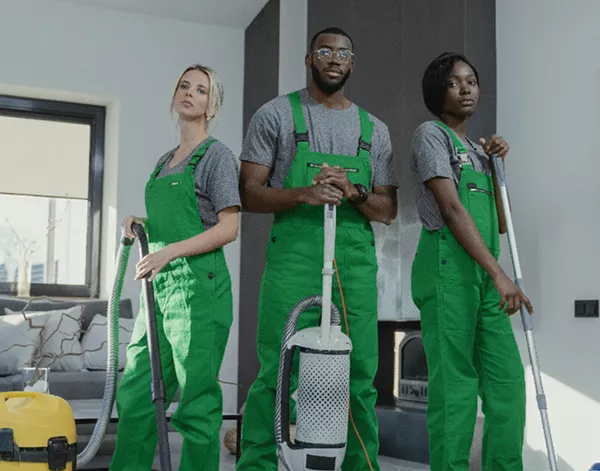Three professional cleaners in bright green overalls standing confidently with cleaning equipment in modern home interior with gray walls
